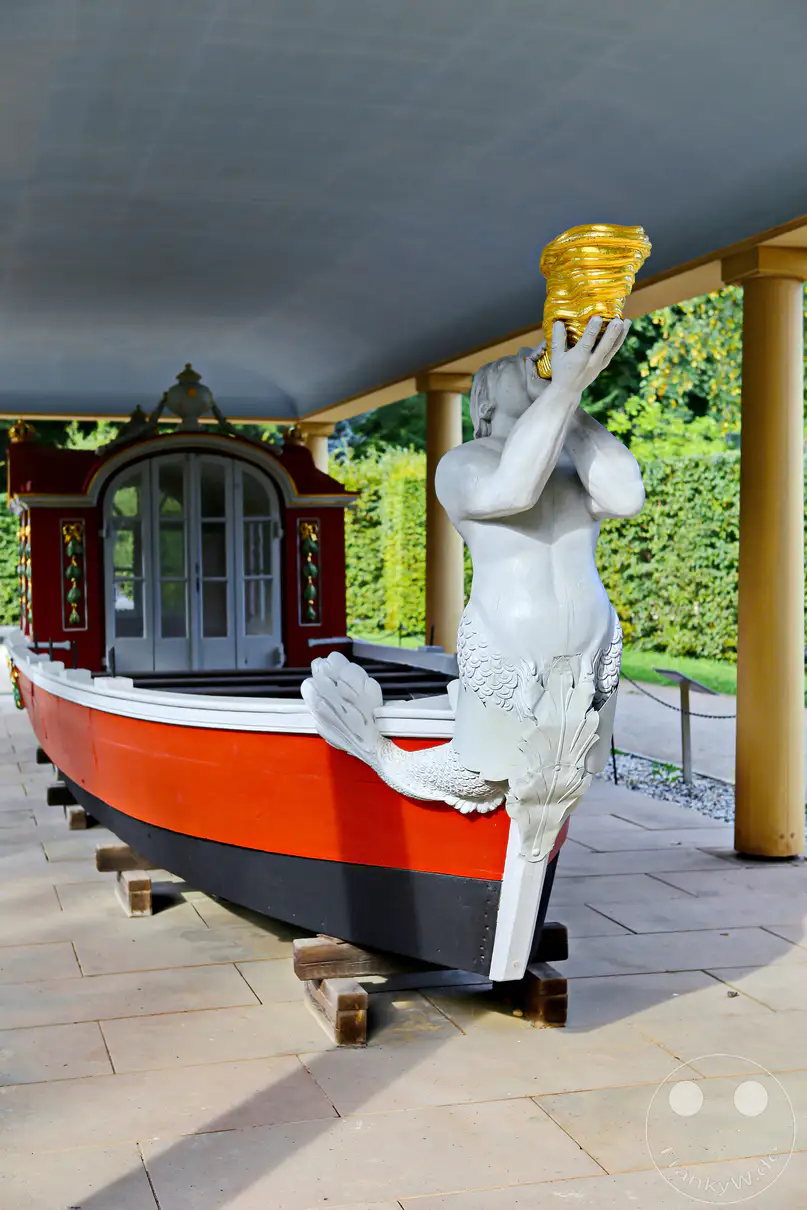 Eine bizarre, weiß bemalte Figur, die einen menschlichen Oberkörper mit einem Fischschwanz kombiniert und einen goldenen Becher hält, ist am Bug einer roten Barkasse platziert. Die kunstvolle Barkasse ruht auf Holzblöcken auf einem gepflasterten Weg, geschützt von einem gelben Säulenportikus im Park der Sommerresidenz Schloss Pillnitz in Deutschland. Hinter der Szenerie erstreckt sich ein üppiger, gepflegter Garten, der eine ruhige und doch märchenhafte Atmosphäre schafft.
