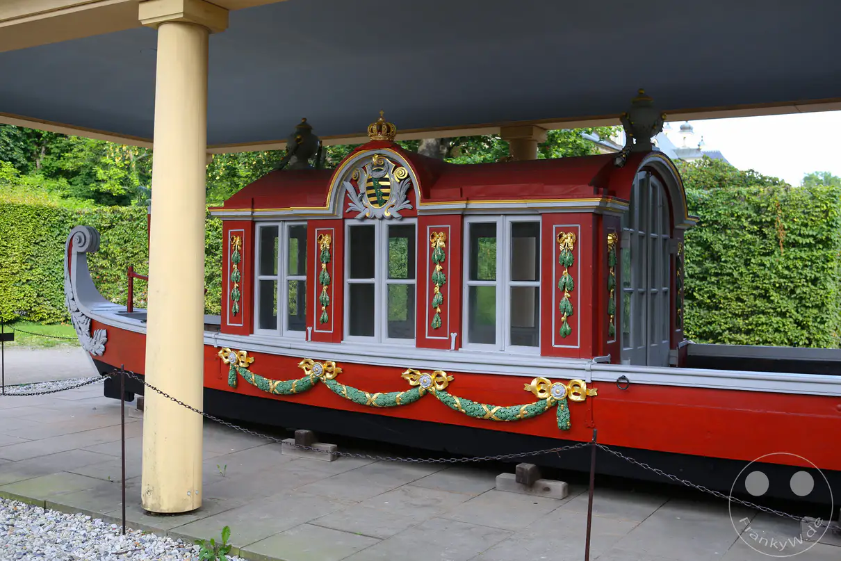 Die rot-schwarz gestrichene Tritonengondel steht geschützt unter einem Pavillondach im Schlossgarten von Pillnitz. Das historische Prunkschiff besitzt einen geschlossenen Kabinenaufbau mit mehreren hellen Fenstern und reich verzierten, goldenen Schnitzereien. Über dem zentralen Kabinenfenster prangt ein aufwendig gestaltetes sächsisches Wappenrelief unter einer goldenen Königskrone. Entlang der Rumpfseite ziehen sich dekorative, grün-goldene Girlandenstrukturen direkt über dem gepflasterten Fundament der Anlage.