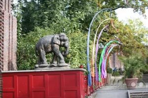 Eine detailreich geschnitzte, dunkle Holzskulptur eines Elefanten thront auf einer roten Brüstung im Außenbereich des Elefanten-Tempels Ganesha Mandir im Leipziger Zoo. Im Hintergrund erstreckt sich der asiatisch gestaltete Tempelgarten, der von hoch aufragenden, bunten Fahnenstangen mit wehenden Bändern, dichten grünen Bäumen und großen Palmenkübeln geschmückt ist. Eine steinerne Buddha-Statue auf einem Sockel sowie das historische Klinkergebäude des Elefantenhauses runden die authentische, fernöstliche Kulisse der weitläufigen Zoo-Anlage ab.