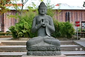 Eine große, aus grauem Naturstein geschlagene Buddha-Statue sitzt in meditativer Lotussitz-Haltung im Außenbereich des Elefanten-Tempels Ganesha Mandir im Leipziger Zoo. Die detaillierte Skulptur thront auf einem verzierten Sockel am Ende einer flachen Steintreppe und formt mit ihren Händen eine klassische Mudra-Geste der Lehre. Der Hintergrund wird von dichten, grünen Bambussträuchern, einem hölzernen Wegweiser zum Elefantenfreigehege und der historischen Klinkerfassade des denkmalgeschützten Elefantenhauses gesäumt, was die fernöstliche Atmosphäre dieses asiatischen Themenbereichs vollendet.