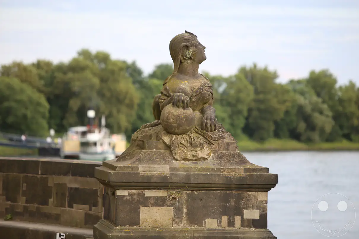 Eine historische Sphinx-Skulptur aus Sandstein thront auf einem massiven Steinpostament direkt am Ufer der Elbe in Pillnitz. Die barocke Figur blickt mit erhobenem Haupt nach oben rechts und hält eine große Kugel unter einer ihrer Tatzen. Auf der rechten Seite erstreckt sich die ruhige Wasseroberfläche des Flusses bis zu den dicht bewaldeten, grünen Hängen am gegenüberliegenden Ufer. Im linken Hintergrund liegt ein historischer Raddampfer der Sächsischen Dampfschifffahrt unscharf am Anlegekai der Schlossanlage.