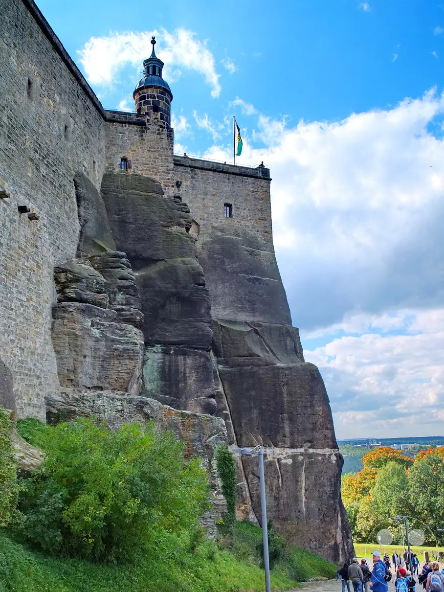 Die Festung Königstein erhebt sich majestätisch auf einem Tafelberg in der Sächsischen Schweiz, Deutschland. Der steile Aufweg und die imposanten Wehrmauern zeugen von ihrer strategischen Bedeutung und Wehrhaftigkeit. Eine sonnige Atmosphäre liegt über der Landschaft, während Besucher die historische Anlage erkunden.