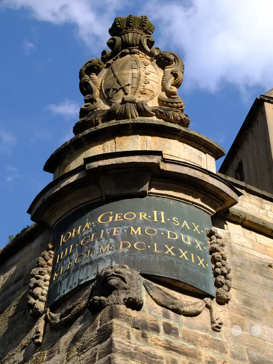 Das detaillierte Wappen von Johann Georg II. von Sachsen schmückt die Fassade der Festung Königstein in der Sächsischen Schweiz. Goldene Lettern auf dunkelgrünem Grund erzählen von der Geschichte dieses monumentalen Bauwerks. Die Nahaufnahme fängt die architektonische Pracht und die Erhabenheit der Festung vor blauem Himmel ein.