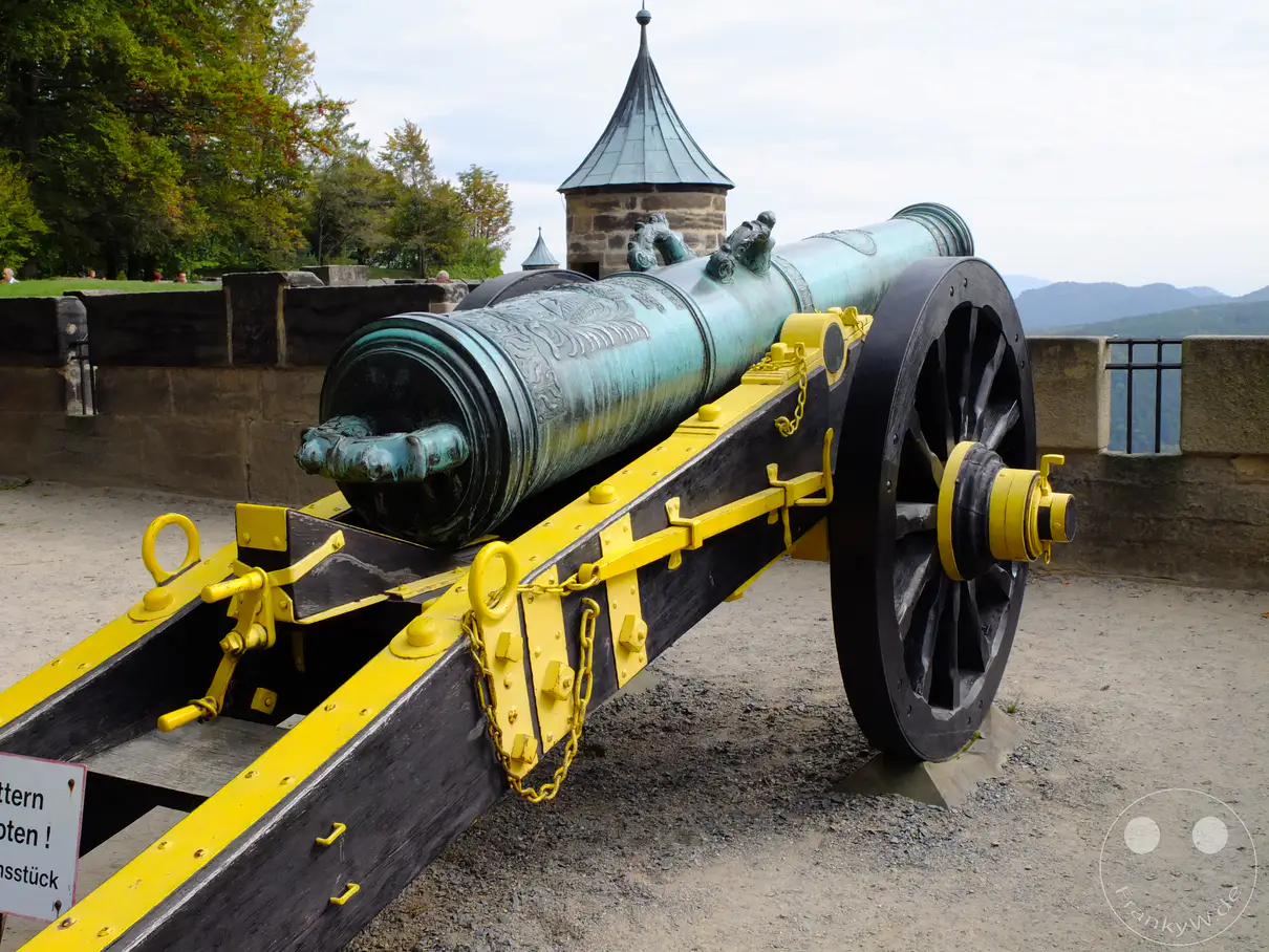Eine imposante historische Belagerungskanone, kunstvoll verziert und auf einer stabilen hölzernen Lafette mit gelben Akzenten, steht auf dem Gelände der Festung Königstein in der Sächsischen Schweiz. Im Hintergrund sind die Festungsmauern und eine Türmchenstruktur zu erkennen, während am Horizont die sanften Hügel der umliegenden Landschaft sichtbar werden. Die Szene vermittelt einen Eindruck von militärischer Geschichte und der erhabenen Atmosphäre dieses bedeutenden deutschen Kulturerbes.