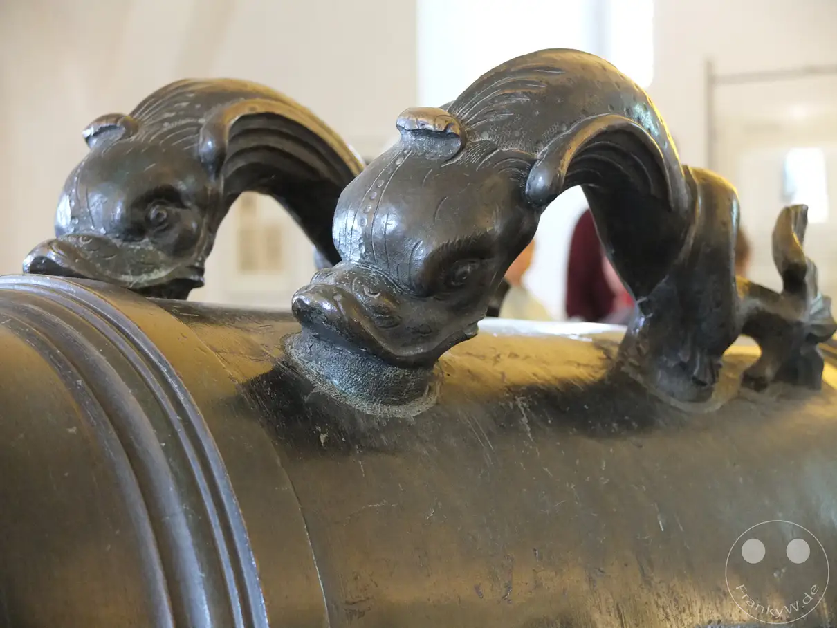 Detailaufnahme der bronzenen Drachenköpfe, die als Henkel einer historischen Kanone im Alten Zeughaus der Festung Königstein in der Sächsischen Schweiz dienen. Die kunstvollen Verzierungen und das massive Material zeugen von vergangener Handwerkskunst und militärischer Bedeutung. Die matte Oberfläche und die detaillierte Ausarbeitung verleihen dem Objekt eine würdevolle und eindrucksvolle Ausstrahlung.