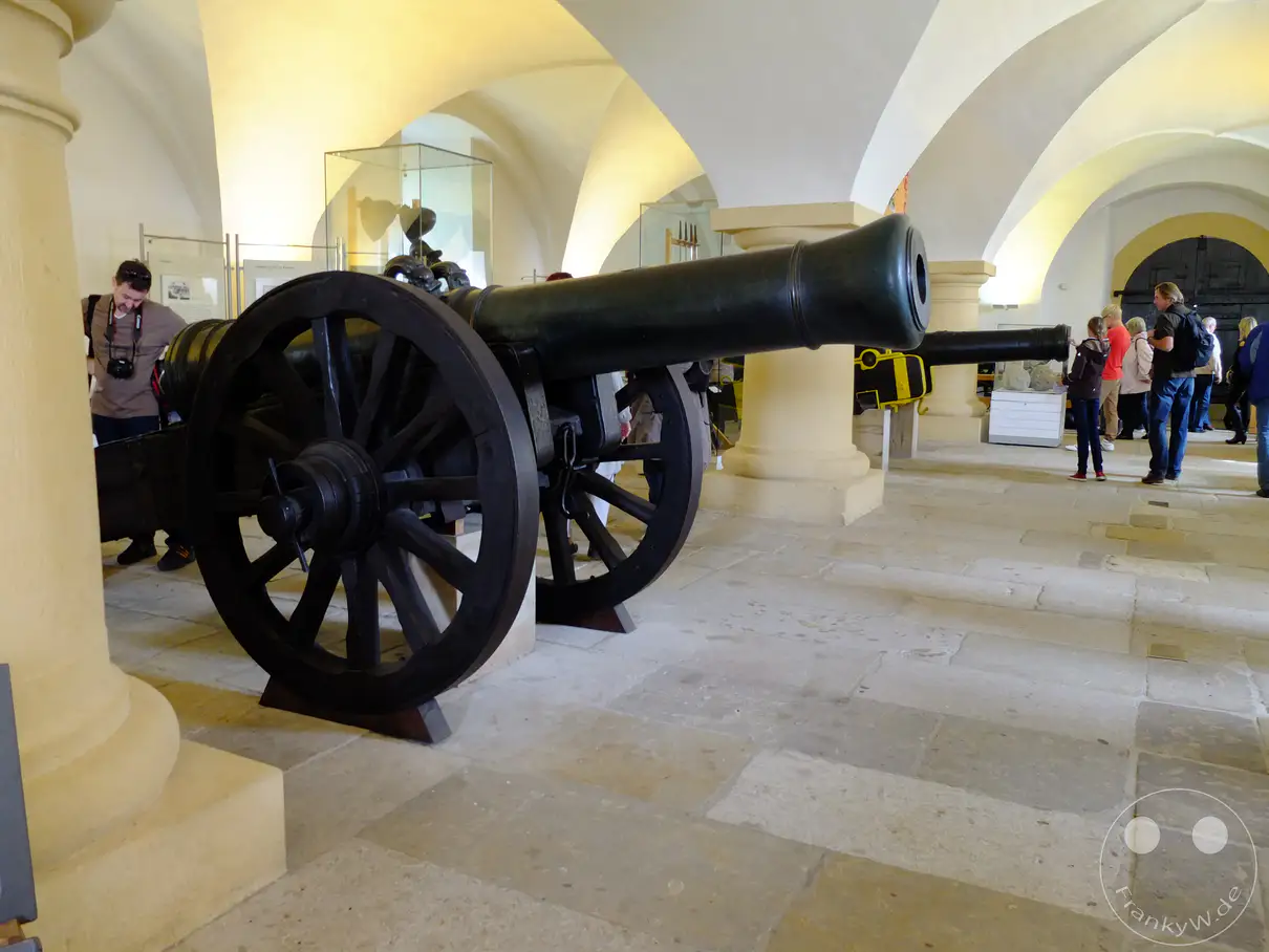 In Deutschland, auf der Festung Königstein in der Sächsischen Schweiz, werden im Alten Zeughaus historische Exponate ausgestellt. Eine imposante Kanone dominiert den Vordergrund des Bildes, während Besucher im Hintergrund durch die Gänge wandeln. Die Atmosphäre ist ruhig und historisch, was die Bedeutung des Ortes unterstreicht.