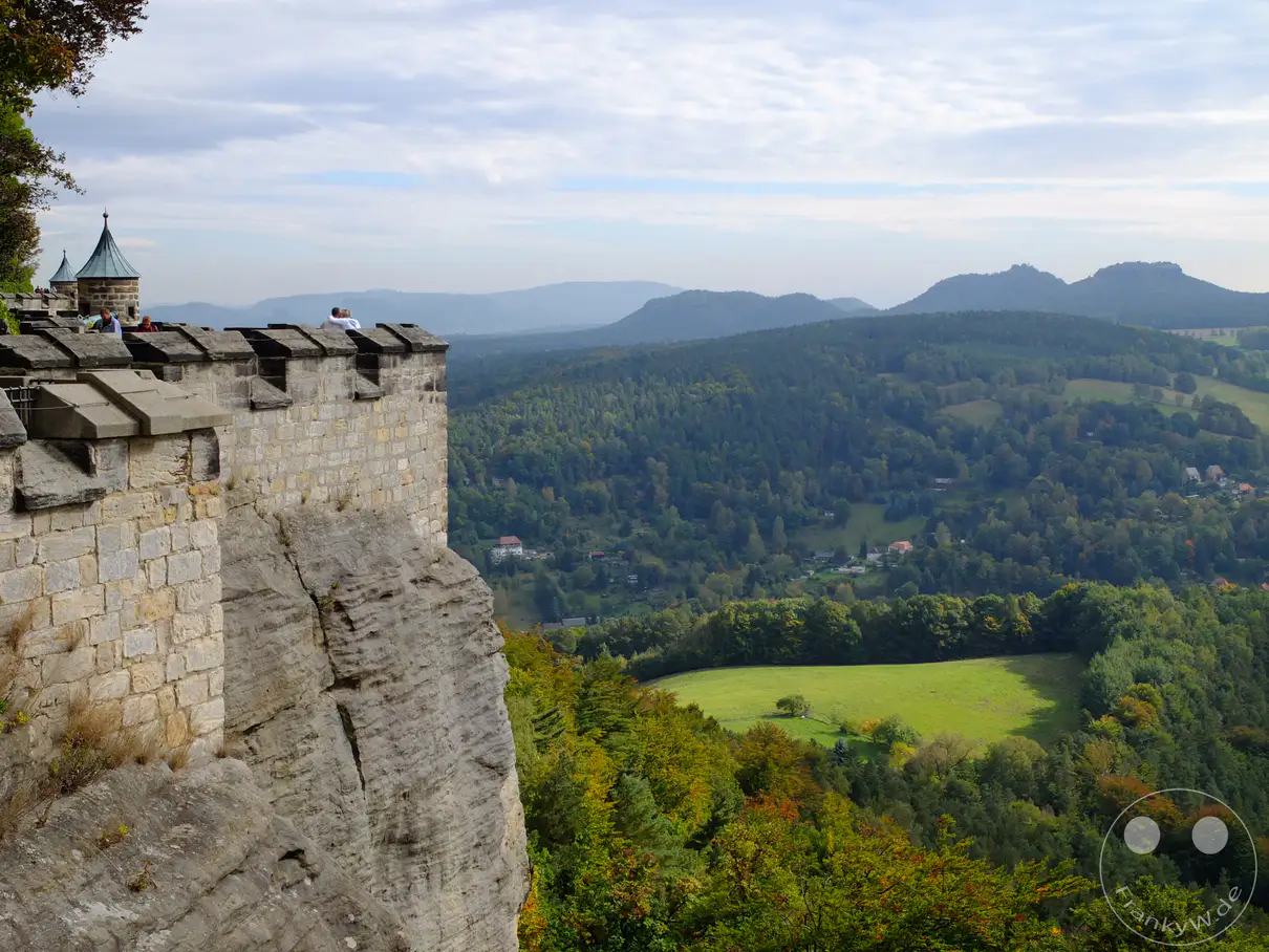 Eine beeindruckende Festungsmauer aus Stein ragt an den Rand einer Felsformation, die in die malerische Landschaft der Sächsischen Schweiz übergeht. Der Blick schweift über tiefgrüne Wälder, sanfte Hügel und vereinzelte Häuser, während sich am Himmel Wolkenballen sammeln. Diese Szenerie strahlt eine friedliche und erhabene Atmosphäre aus, die von der historischen Festung Königstein überragt wird.