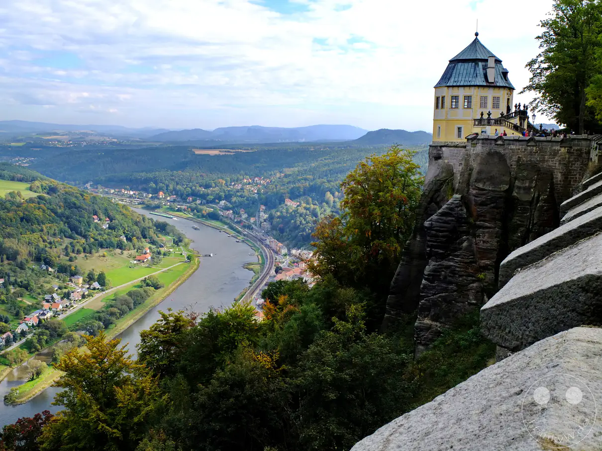 Ein weitläufiger Blick von der Festung Königstein in der Sächsischen Schweiz offenbart die majestätische Elbe, die sich durch eine grüne Flusslandschaft schlängelt. Auf der rechten Seite thront die Friedrichsburg auf einem Felsen, während sich im Tal charmante Dörfer und üppige Wälder erstrecken. Die Szenerie versprüht eine friedliche und erhabene Atmosphäre, die von der Schönheit der Natur und der historischen Bedeutung des Ortes geprägt ist.