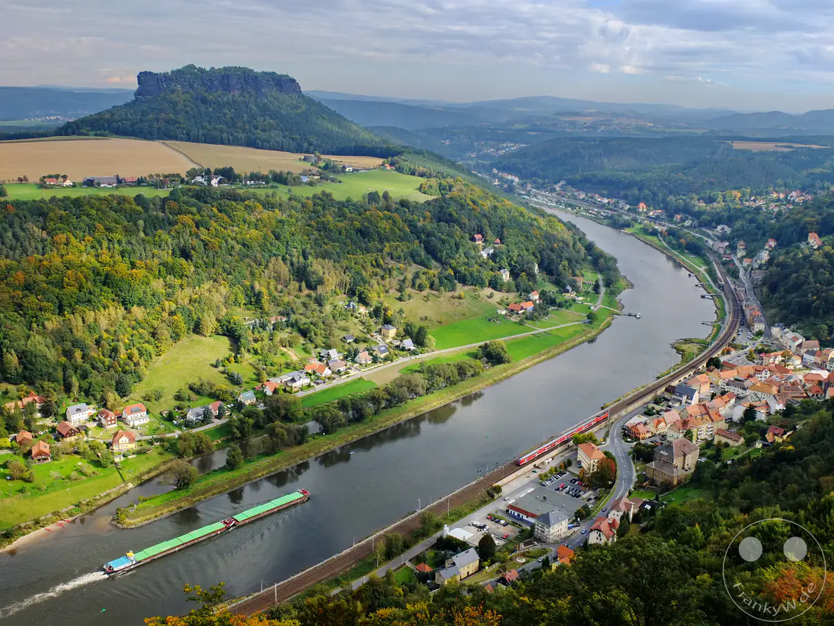 Ein majestätischer Ausblick auf die Sächsische Schweiz in Deutschland, wo sich die Elbe malerisch durch die Landschaft schlängelt. Die imposante Festung Königstein thront über dem Elbtal, während im Vordergrund ein Frachtschiff auf dem Fluss unterwegs ist und sich ein kleiner Ort am Ufer schmiegt. Die Szenerie strahlt eine friedliche und erhabene Atmosphäre aus, die die natürliche Schönheit und die reiche Geschichte der Region widerspiegelt.