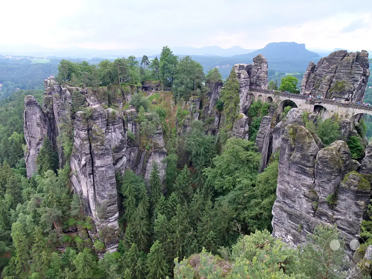 Ein Blick über die majestätische Basteibrücke und die zerklüfteten Sandsteinfelsen der Sächsischen Schweiz. Die imposante Steinbrücke verbindet zwei Felsengruppen und bietet atemberaubende Ausblicke auf die bewaldete Landschaft und das Elbtal. Die Szenerie strahlt eine beeindruckende Naturkulisse und historische Architektur aus.