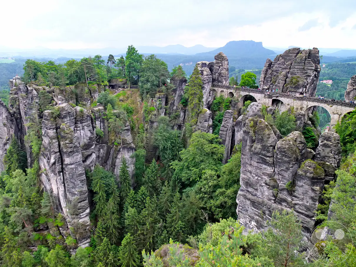 Die berühmte Basteibrücke überspannt schroff aufragende Sandsteinfelsen in der Sächsischen Schweiz, Deutschland. Touristen bevölkern den steinernen Viadukt und genießen den atemberaubenden Blick über das Elbtal und die umliegende Berglandschaft. Die Szene strahlt eine majestätische Ruhe und die Erhabenheit der Natur aus.