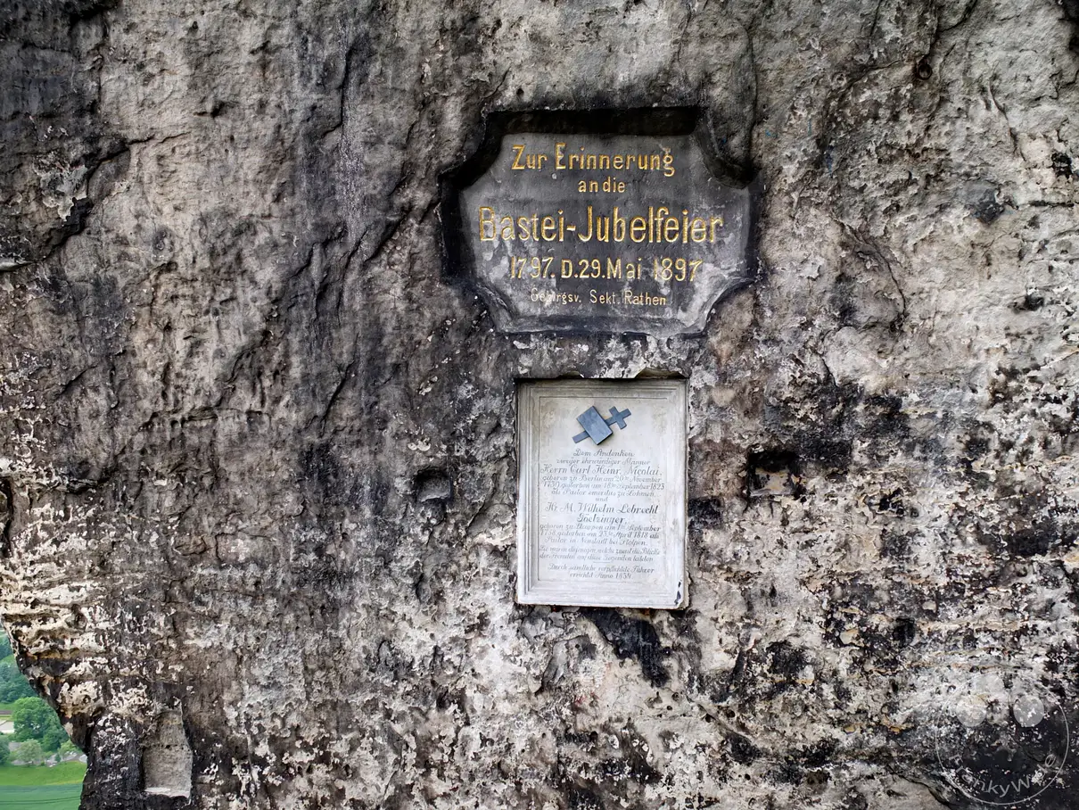 Eine Gedenktafel ist auf einem rauen Felsen in der Sächsischen Schweiz angebracht. Goldene Schrift erinnert an die Bastei-Jubelfeier und zwei Gedenkpersonen. Die Inschrift ist in die steinerne Oberfläche eines Felsens gemeißelt und strahlt eine historische und ehrwürdige Stimmung aus.