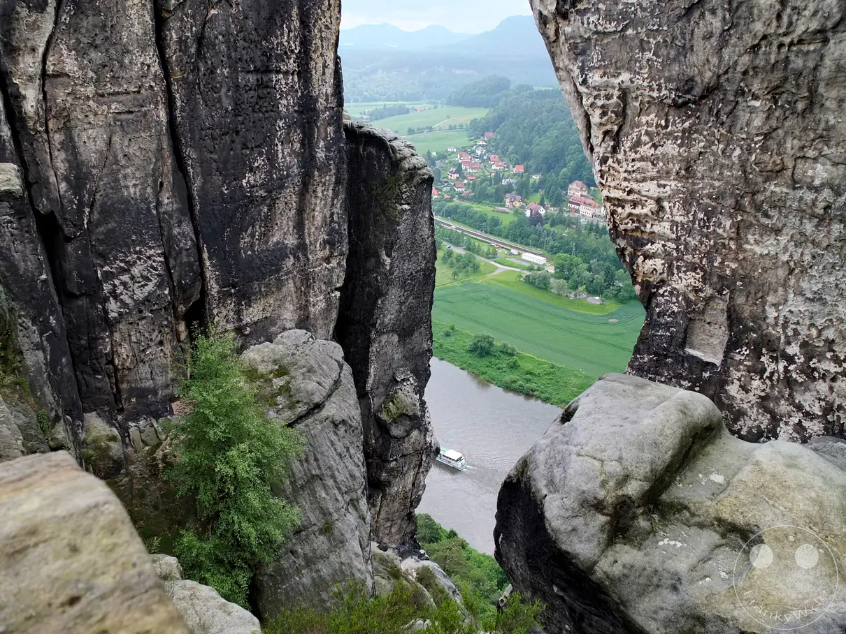 Zu sehen ist der Tiefblick durch ein schmales Felsentor im Basteigebiet hinab auf die Elbe. Mächtige, dunkel verwitterte Sandsteinwände rahmen den Blick auf das Flusstal und ein vorbeifahrendes Ausflugsschiff ein. Im Hintergrund erstrecken sich die grünen Wiesen, Felder und Häuser des Kurorts Rathen entlang des Flussufers. Ein kleiner, grüner Baum krallt sich direkt am Abgrund in die zerklüfteten Felsen im Vordergrund.