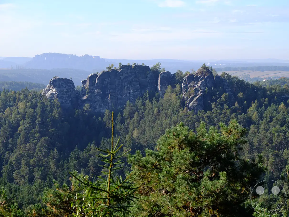 Ein weitläufiger Panoramablick über das Elbsandsteingebirge in der Sächsischen Schweiz offenbart dramatische Sandsteinfelsformationen, die von dichten Nadelwäldern umgeben sind. Die Szenerie wird von einem klaren Himmel mit vereinzelten Wolken überragt, was eine friedliche und erhabene Atmosphäre schafft. In der Ferne deuten sich sanfte Hügel und ländliche Gebiete an, die die Weite der Landschaft unterstreichen.