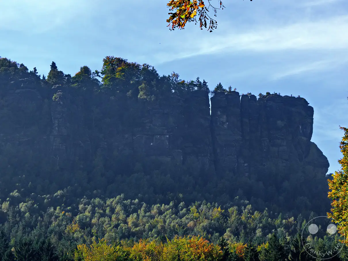 Die imposanten Felsformationen der Sächsischen Schweiz ragen dramatisch in den blauen Himmel. Ein tiefer Felsspalt durchzieht die massiven Sandsteinwände, über die sich in der Ferne eine Schluchtbrücke spannt und die Weite der Landschaft unterstreicht. Bunte Herbstbäume säumen den Fuß der Felsen und schaffen einen farbenfrohen Kontrast zur schroffen Naturformation in Deutschland.