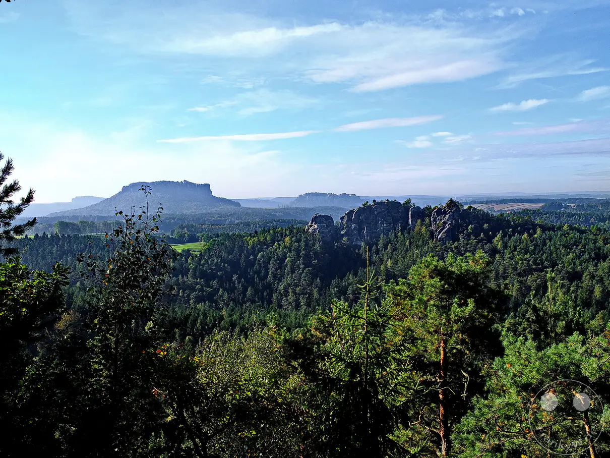 Ein weitläufiger Panoramablick erstreckt sich über das Elbsandsteingebirge in der Sächsischen Schweiz, Deutschland. Grüne Wälder bedecken die Hügel, während markante Sandsteinfelsen, wie der Rauenstein, majestätisch emporragen und die bergige Landschaft prägen. Der Himmel ist blau mit vereinzelten Wolken, was eine friedliche und erhabene Atmosphäre schafft.