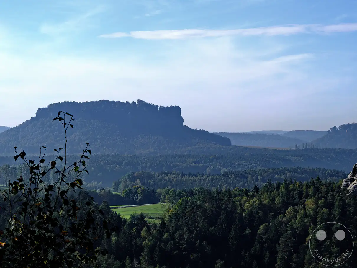 Ein dunstiger Blick über das Elbsandsteingebirge in der Sächsischen Schweiz, Deutschland, enthüllt die markante Silhouette des Liliensteins. Dichte Wälder bedecken die sanften Hänge und gehen in der Ferne in weitere Hügelketten über, die in der Morgensonne bläulich schimmern. Vor dem Vordergrund ragen die silhouettenhaften Zweige eines Baumes auf und verleihen der weiten Landschaft eine zusätzliche Tiefe.