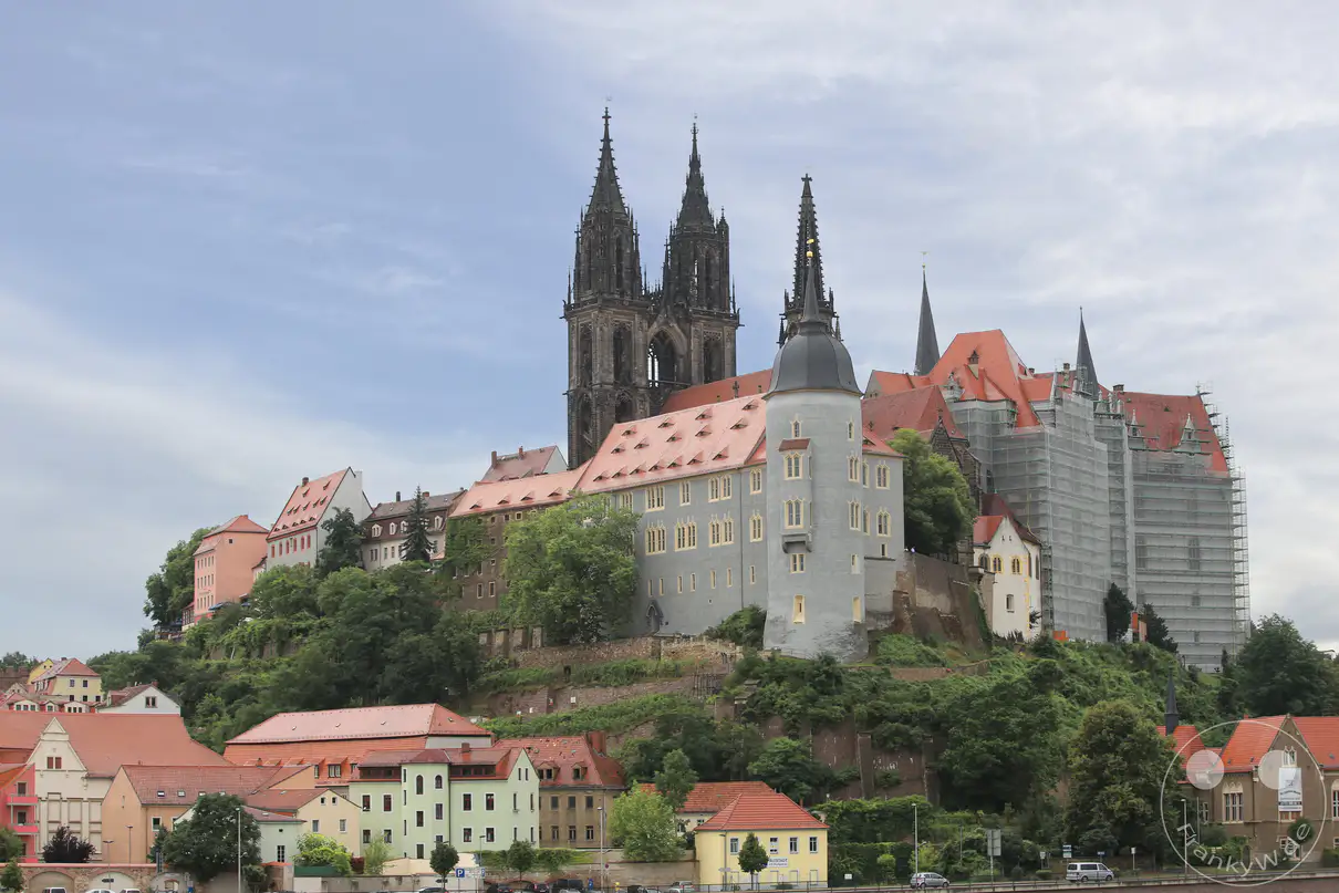 Das monumentale Ensemble aus Albrechtsburg und Meißner Dom thront auf dem grünen Burgberg über den Dächern der sächsischen Stadt Meißen. Die gotische Architektur des Doms zeichnet sich durch zwei hohe, filigrane Turmspitzen aus dunklem Stein im Zentrum der Anlage aus. Der weitläufige Schlosskomplex erstreckt sich mit hellen Fassaden, einem runden Eckturm und großen, rötlich gedeckten Satteldächern entlang des Plateaus. Am rechten Bildrand umgibt ein großes Baugerüst einen Teil der historischen Mauern, während im Vordergrund mehrstöckige Wohnhäuser den Fuß des Hanges säumen.