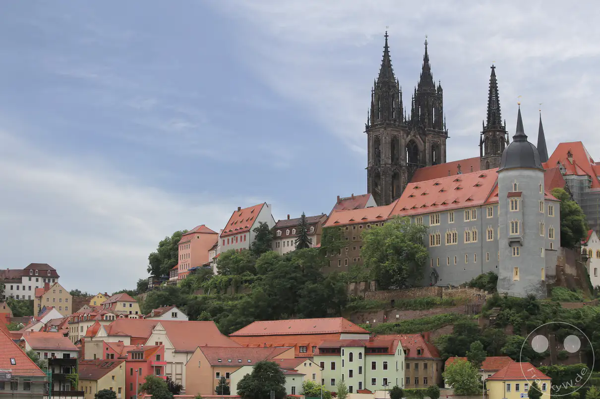 Das monumentale Ensemble aus Albrechtsburg und Meißner Dom thront auf dem grünen Burgberg über den Dächern der sächsischen Stadt Meißen. Die gotische Architektur des Doms zeichnet sich durch zwei hohe, filigrane Turmspitzen aus dunklem Stein im Zentrum der Anlage aus. Der weitläufige Schlosskomplex erstreckt sich mit hellen Fassaden, einem runden Eckturm und großen, rötlich gedeckten Satteldächern entlang des Plateaus. Am rechten Bildrand umgibt ein großes Baugerüst einen Teil der historischen Mauern, während im Vordergrund mehrstöckige Wohnhäuser den Fuß des Hanges säumen.