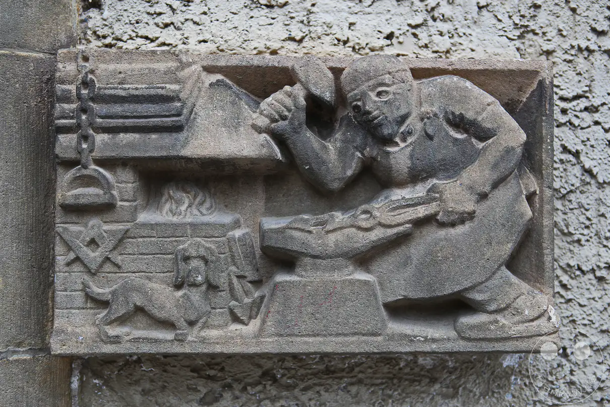 Ein detailreiches, historisches Steinrelief ist an einer rau verputzten Fassadenwand des Stadtmuseums in Meißen angebracht. Das plastisch ausgearbeitete Motiv zeigt einen kräftigen Schmied mit erhobenem Hammer bei der Arbeit an einem Amboss. Auf der linken Seite befindet sich eine gemauerte Herdstelle mit züngelnden Flammen, einer Kettensuspension und einem kleinen Hund im Vordergrund. Die gesamte sandsteinartige Skulptur fügt sich als dekoratives Bauelement direkt in das historische Mauerwerk der Außenwand ein.