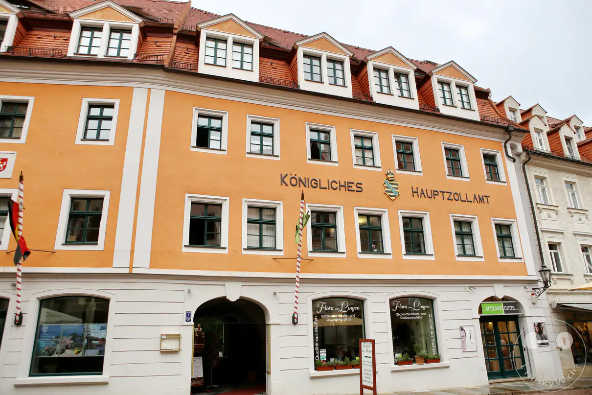 Die markante orangefarbene Fassade des Königlichen Hauptzollamtes in Meißen erstreckt sich unter einem bedeckten Himmel. Die goldene Aufschrift des historischen Behördennamens prangt im Zentrum des Gebäudes direkt über einer Reihe von rechteckigen Fenstern. Das mehrstöckige Bauwerk besitzt ein großes, rötlich gedecktes Mansarddach mit zahlreichen weißen Dachgauben in zwei Ebenen. Im weiß verputzten Erdgeschoss befinden sich mehrere Ladengeschäfte mit Schaufenstern sowie ein großes Rundbogenportal als Haupteingang.