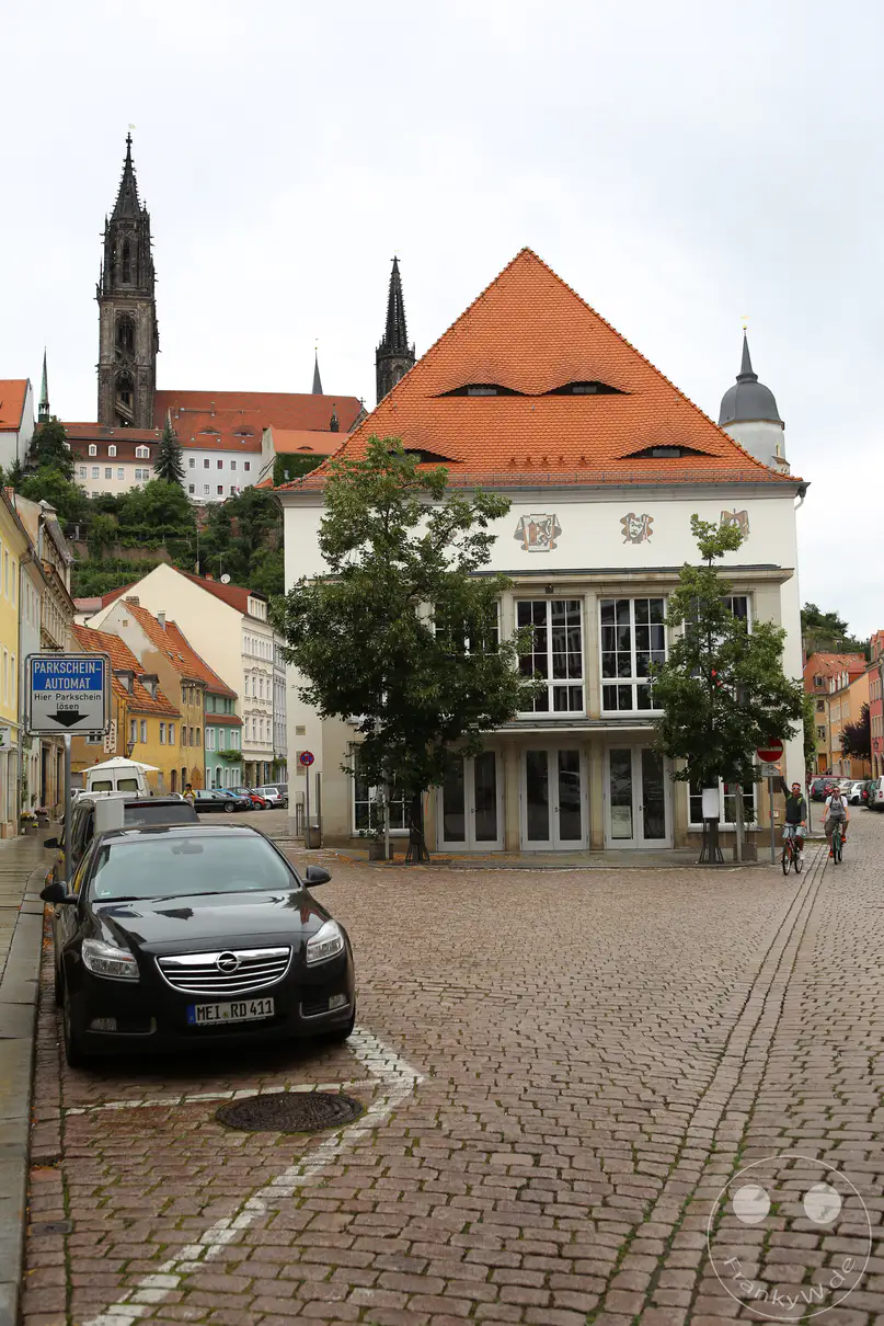 Das hell verputzte Theatergebäude in Meißen steht an einem gepflasterten Platz unter einem wolkenverhangenen Himmel. Ein markantes, steiles Satteldach mit mehreren Hechtgauben prägt die obere Architektur des historischen Kulturbauwerks. Im Hintergrund ragen die markanten Türme der Frauenkirche und des Doms auf dem Burgberg über die umliegenden Häuserzeilen empor. Ein schwarzer Pkw parkt im linken Vordergrund auf dem Kopfsteinpflaster nahe einem Verkehrsschild für Parkscheine.