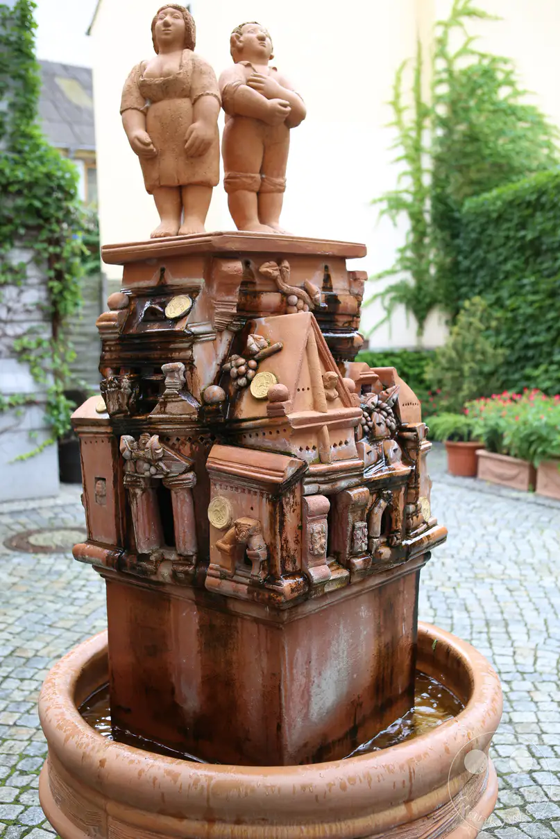 Ein mehrstufiger, rötlicher Tonbrunnen steht im Zentrum eines gepflasterten Innenhofs im ehemaligen Zollhof in Meißen. Auf der Spitze des architektonisch gestalteten Keramikaufbaus thronen zwei menschliche Figuren in Form einer Frau und eines Mannes. Der Brunnenkörper imitiert historische Hausfassaden mit kleinen Säulen, Giebeln und eingelassenen goldenen Münzen im fließenden Wasser. Ein kreisrundes, ebenfalls aus Ton gefertigtes Auffangbecken bildet die Basis der nassen Skulptur vor einem Hintergrund aus grünen Kletterpflanzen.