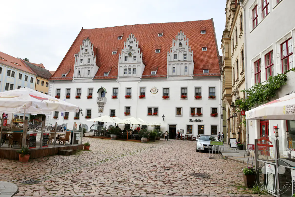 Die weiß verputzte Fassade des spätgotischen Rathauses in Meißen erstreckt sich hinter einem weitläufigen Pflasterplatz unter einem bewölkten Himmel. Drei prachtvolle, reich verzierte Ziergiebel ragen symmetrisch aus dem großen, rötlich gedeckten Satteldach empor. Rote Blumenkästen schmücken die zahlreichen Fensterreihen des historischen Verwaltungsgebäudes über der gesamten Front. Im Erdgeschoss laden die hellen Sonnenschirme und Tische eines Außencafés vor den steinernen Portalen zum Verweilen ein.