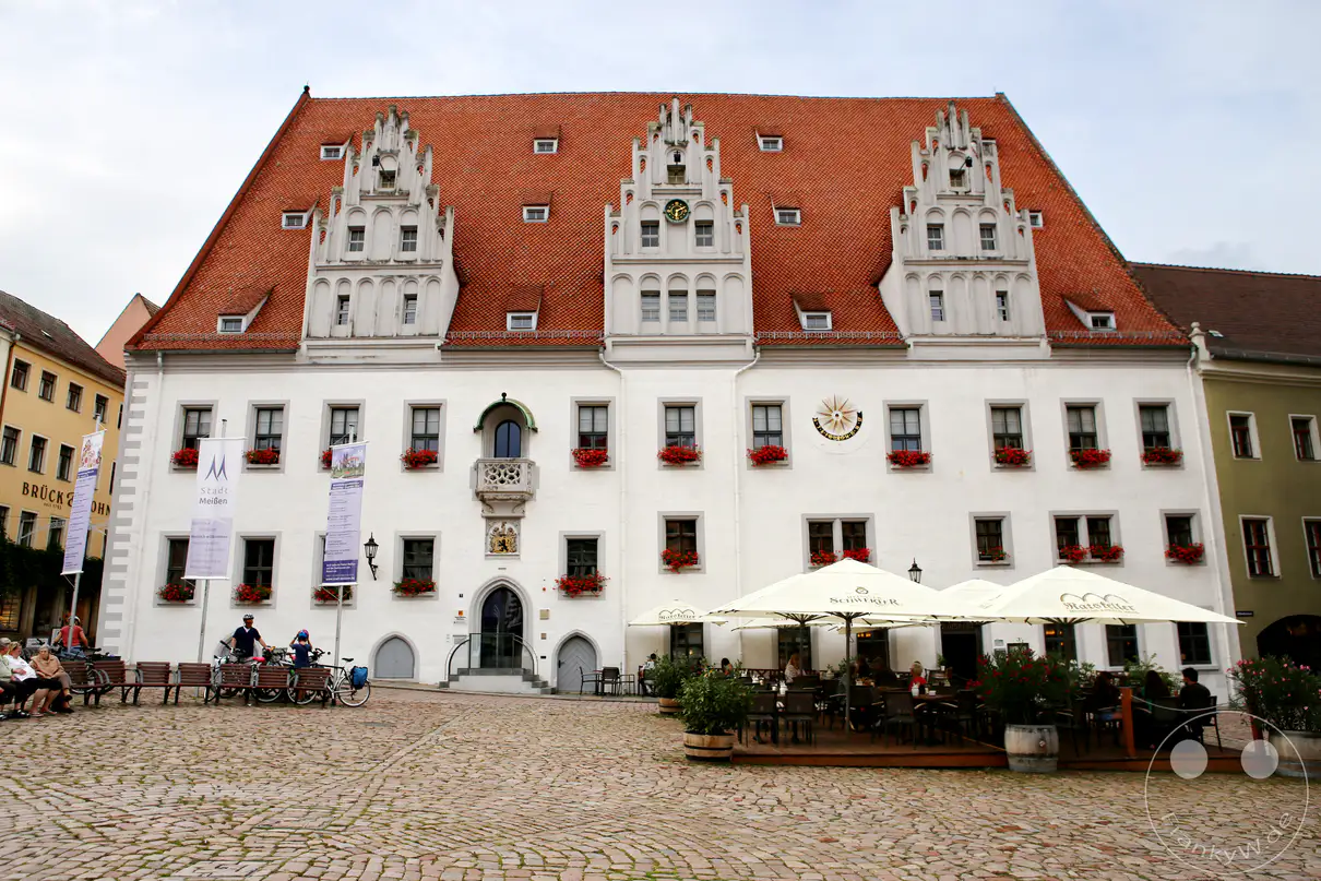 Die weiß verputzte Fassade des spätgotischen Rathauses in Meißen erstreckt sich hinter einem weitläufigen Pflasterplatz unter einem bewölkten Himmel. Drei prachtvolle, reich verzierte Ziergiebel ragen symmetrisch aus dem großen, rötlich gedeckten Satteldach empor. Rote Blumenkästen schmücken die zahlreichen Fensterreihen des historischen Verwaltungsgebäudes über der gesamten Front. Im Erdgeschoss laden die hellen Sonnenschirme und Tische eines Außencafés vor den steinernen Portalen zum Verweilen ein.