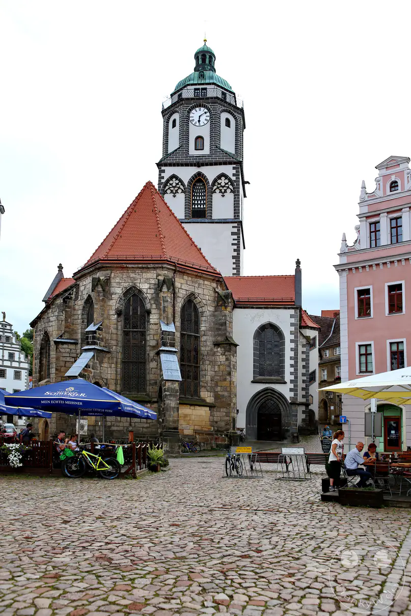 Die historische Frauenkirche in Meißen steht mit ihrem markanten, weiß-schwarz verzierten Turm an einem gepflasterten Platz unter einem hellen Himmel. Der gotische Chorraum des Sakralbaus ist aus unregelmäßigen, dunklen Natursteinen errichtet und mit einem rötlichen Zeltdach gedeckt. Im Vordergrund erstreckt sich eine Außengastronomie mit großen blauen Sonnenschirmen und abgeparkten Fahrrädern auf dem weitläufigen Kopfsteinpflaster. Mehrstöckige, farbenfrohe Bürgerhäuser mit verzierten Giebeln rahmen die Szenerie auf der linken und rechten Bildseite ein.