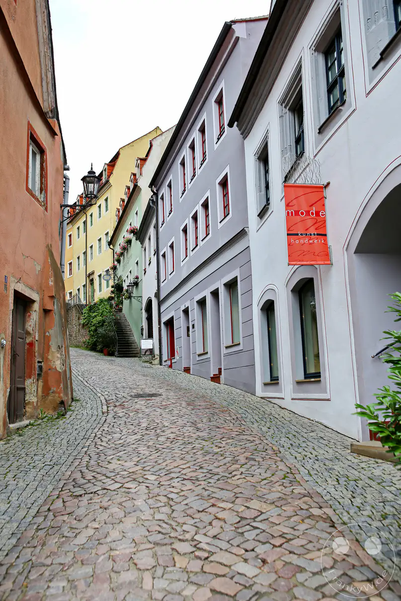 Eine charmante Gasse in Meißen, Deutschland, schlängelt sich bergauf, gesäumt von historischen Gebäuden mit bunten Fassaden. Das Kopfsteinpflaster und die traditionelle Architektur erzeugen eine malerische und zeitlose Atmosphäre in der Altstadt. Ein kleiner Laden mit der Aufschrift "mode kunst handwerk" deutet auf lokale Handwerkskunst und Stil hin, während die verträumte Kulisse zum Erkunden einlädt.