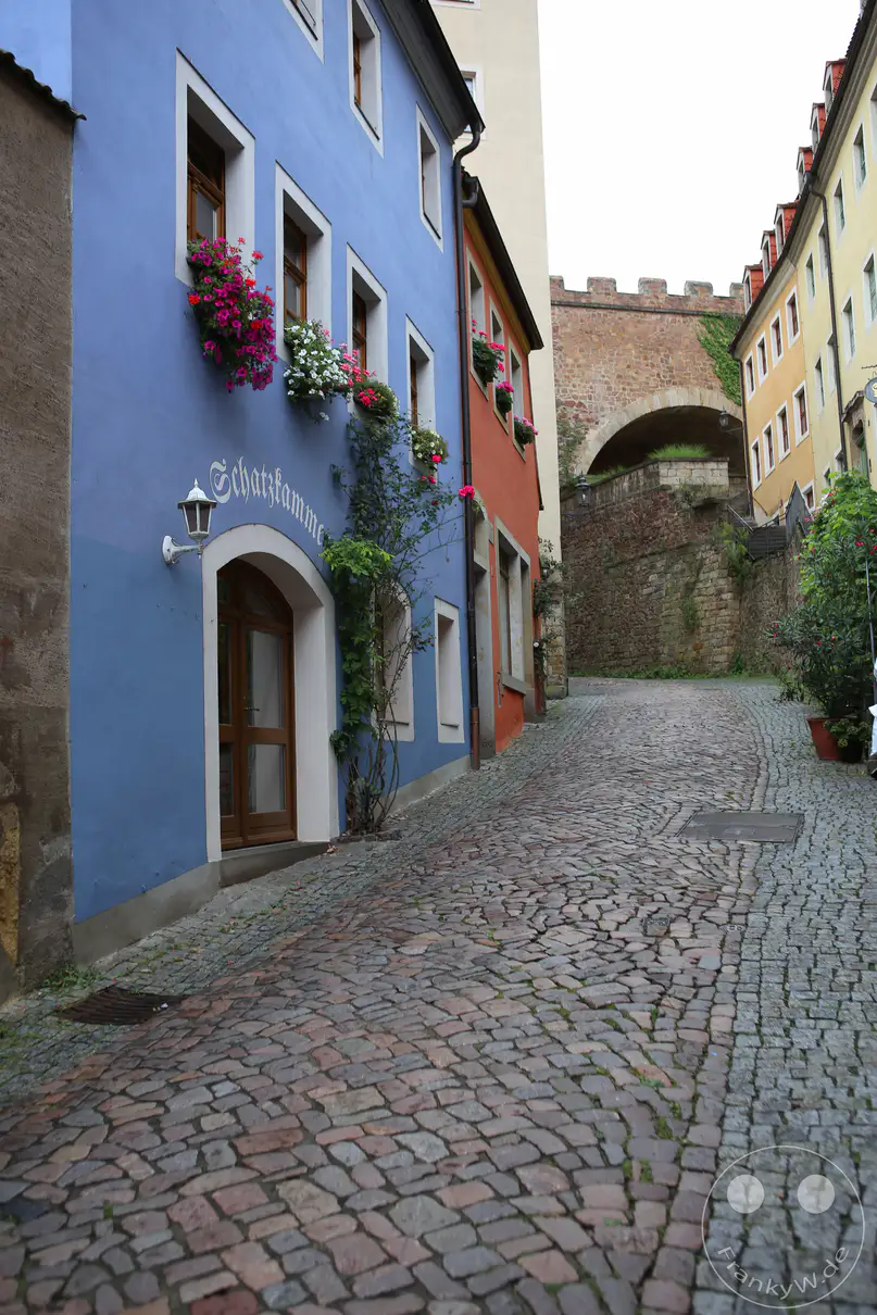 Die schmale, ansteigende Gasse des Hohlwegs schlängelt sich mit historischem Kopfsteinpflaster durch die Altstadt von Meißen. Auf der linken Seite zieht eine auffällig blau verputzte Gebäudefassade mit der geschwungenen Aufschrift „Schatzkammer“ über dem hölzernen Portal die Blicke auf sich. Farbenfroher Blumenschmuck in den Fensterkästen und rankendes Grün beleben das historische Straßenbild. Am oberen Ende des Weges überspannt ein massiver steinerner Torbogen der Schlossstufen die Szenerie vor den Mauern des Burgbergs.