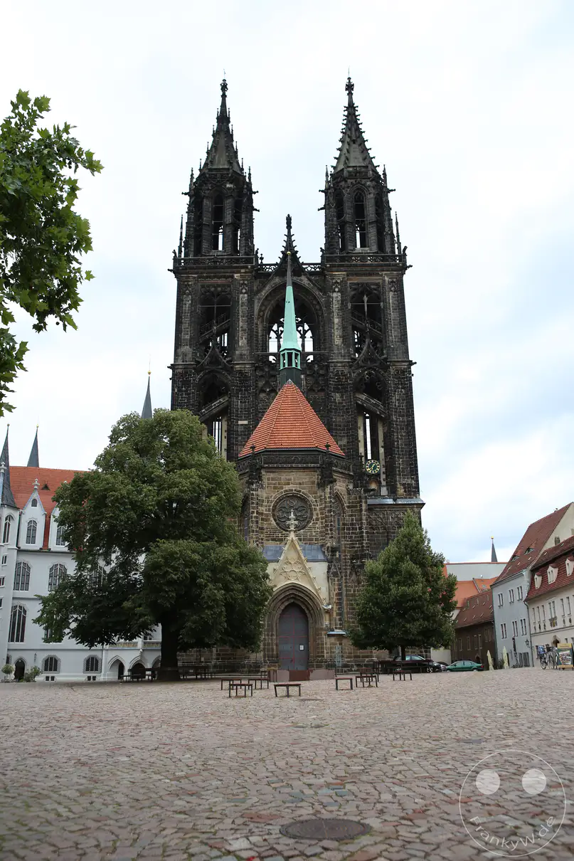 Die monumentale Westfassade des Meißner Doms erhebt sich hinter einem weitläufigen, gepflasterten Platz in der Altstadt von Meißen. Zwei hohe, filigran durchbrochene Türme aus dunklem Naturstein ragen symmetrisch in den wolkenverhangenen Himmel. Vor dem gotischen Hauptbaukörper steht die Johanneskapelle mit einem rötlichen Zeltdach und einer schlanken, grünlichen Turmspitze. Zwei große, dichte Laubbäume und vereinzelte Holzbänke flankieren den gepflasterten Domplatz im Vordergrund der sakralen Anlage.