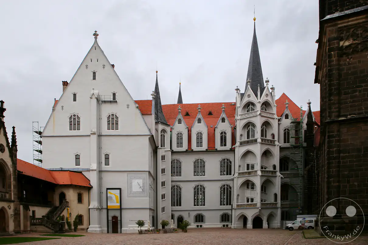 Die hell verputzten Fassaden der Albrechtsburg in Meißen erstrecken sich hinter dem gepflasterten Domplatz unter einem bewölkten Himmel. Im Zentrum des historischen Schlosskomplexes ragt der Große Wendelstein als kunstvoller, reich verzierter Treppenturm mit einer spitzen dunklen Haube empor. Große gotische Vorhangbogenfenster und steile, rötlich gedeckte Satteldächer prägen die Architektur des spätgotischen Residenzschlosses. Am rechten Bildrand schließt die massive, dunkle Natursteinwand des Meißner Doms die Szenerie ab.
