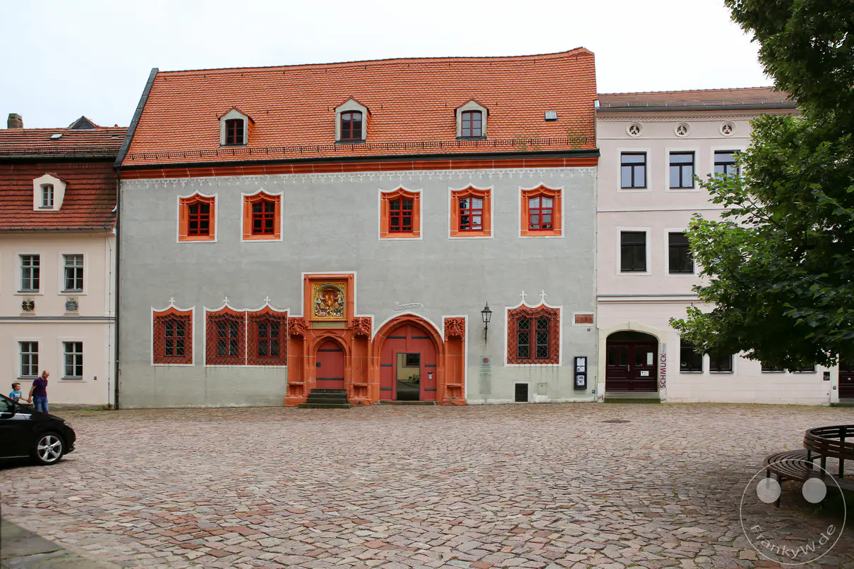 Die graue Fassade des historischen Bischofsschlosses am Domplatz in Meißen steht unter einem bewölkten Himmel. Ein prachtvolles, rötlich gestaltetes Renaissance-Portal mit einem farbigen Wappenrelief bildet den zentralen Zugang im Erdgeschoss des Gebäudes. Ein großes, ziegelgedecktes Satteldach mit drei symmetrisch angeordneten Dachgauben schließt den historischen Bau nach oben ab. Vor dem Bauwerk erstreckt sich das weitläufige Kopfsteinpflaster des Platzes, das auf der rechten Seite von einer halbrunden Sitzbank und dichten Baumkronen flankiert wird.