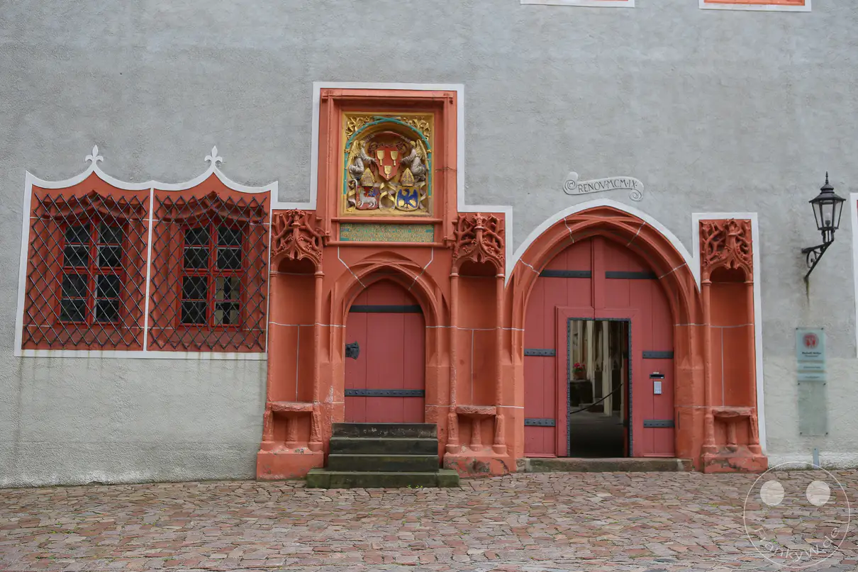 Das prachtvoll gestaltete, rötliche Sandsteinportal des Bischofsschlosses am Domplatz in Meißen bildet das zentrale Element der grau verputzten Erdgeschossfassade. Ein aufwendig ausgearbeitetes, farbiges Wappenrelief mit goldenen Akzenten thront direkt über der kleineren, linken Holztür mit vorgelagerten Steinstufen. Eine größere, rundbogige Toreinfahrt auf der rechten Seite besitzt eine integrierte, geöffnete Schlupftür mit Blick in den Innenraum. Links flankieren zwei hohe, von kunstvoll geschmiedeten roten Gittern geschützte Fenster den historischen Eingangsbereich oberhalb des unregelmäßigen Kopfsteinpflasters.