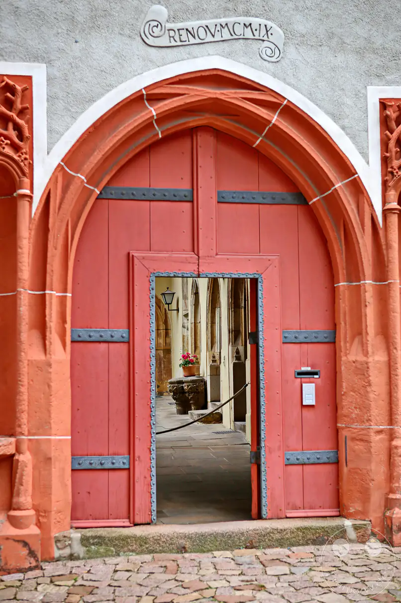 Das Bild zeigt ein imposantes, rotes, gotisches Holztor, das zu einem Innenhof führt. Das Tor ist mit Metallbeschlägen verziert und über dem Bogen ist die Jahreszahl "RENOV MCM IX" (1909) eingraviert. Die Szene strahlt eine historische und ehrwürdige Atmosphäre aus, ein Blick in eine vergangene Zeit in Meißen.