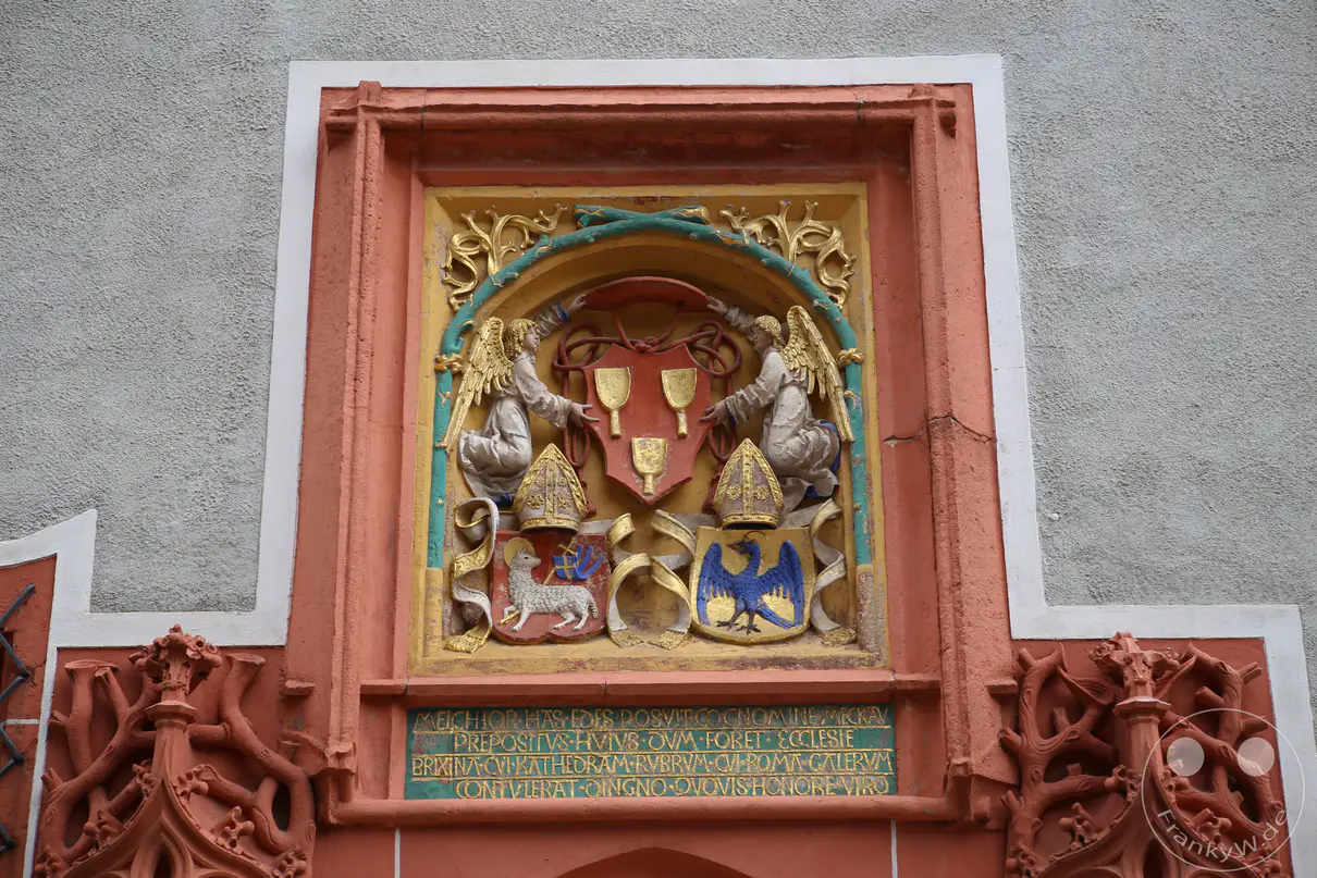 Eine Detailaufnahme zeigt das farbenprächtige Wappenrelief des Melchior von Meckau über dem Portal des Bischofsschlosses in Meißen. Zwei plastisch ausgeformte Engel mit goldenen Flügeln halten das zentrale, rote Schildmotiv mit drei Kelchen im oberen Drittel der steinernen Tafel. Darunter befinden sich zwei kleinere Wappenschilde mit Darstellungen eines Lammes sowie eines blauen Adlers, jeweils von einer goldenen Mitra bekrönt. Eine mehrzeilige, in den rötlichen Sandstein gemeißelte lateinische Inschrift schließt das historische Kunstwerk an der grau verputzten Fassade nach unten hin ab.