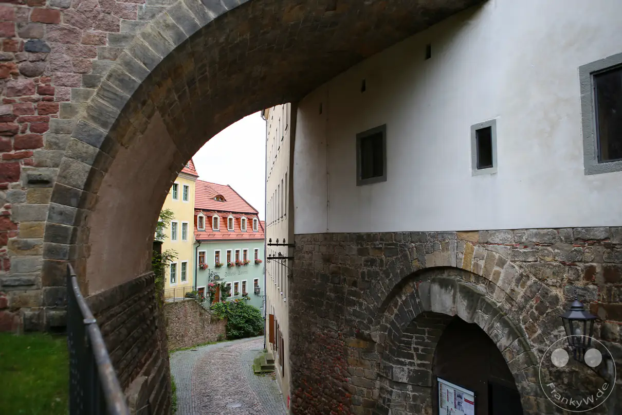 Ein massiver, steinerner Torbogen überspannt den ansteigenden Pflasterweg des Hohlwegs in Meißen. Auf der rechten Seite schließt sich eine historische Bruchsteinmauer mit einem weiteren, kleineren Rundbogenportal und einer klassischen Wandlaterne an das Bauwerk an. Durch den großen Hauptbogen hindurch öffnet sich der Blick auf eine schmale Gasse mit mehrstöckigen, farbenfrohen Wohnhäusern unter rötlichen Ziegeldächern. Eine rustikale, gemauerte Befestigungswand flankiert den gepflasterten Verbindungsweg zur Albrechtsburg auf der linken Seite.