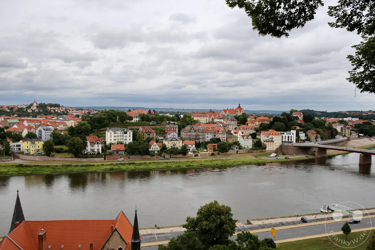 Ein weiter Panoramablick aus einer erhöhten Perspektive zeigt die Stadt Meißen und den Verlauf der Elbe unter einem dicht bewölkten Himmel. Der breite Fluss zieht sich horizontal durch die Mitte des Bildes und trennt das grüne Ufer im Vordergrund von den bebauten Hängen auf der gegenüberliegenden Seite. Zahlreiche mehrstöckige Wohnhäuser und Villen mit roten Ziegeldächern staffeln sich entlang der Hügelkette im Hintergrund. Eine moderne Brücke überspannt das Gewässer auf der rechten Seite, während dichte Baumkronen die Szenerie am oberen rechten Rand natürlich einrahmen.