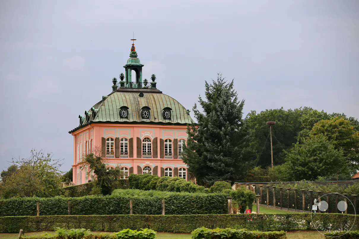Das Fasanenschlösschen in Moritzburg, Sachsen, präsentiert sich in pastellfarbenem Barockstil inmitten einer gepflegten Gartenanlage. Die geschwungene Dachform und die detaillierte Architektur des Lustschlosses strahlen eine elegante und fast märchenhafte Atmosphäre aus. Umgeben von üppigem Grün und bewölktem Himmel, lädt die Szenerie zu einem Besuch in eine vergangene Epoche ein.
