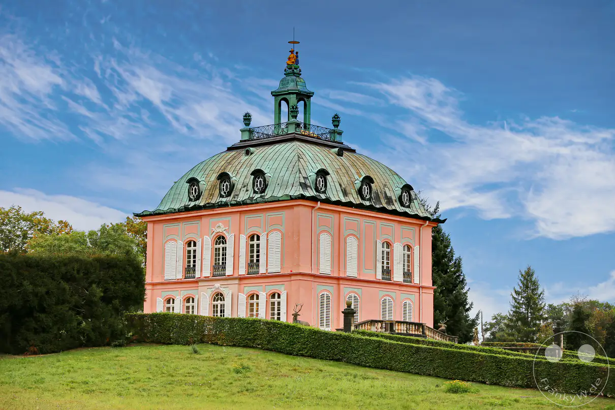 Das barocke Fasanenschlösschen in Moritzburg, Deutschland, erhebt sich prunkvoll mit seiner geschwungenen Kupferkuppel und der rosafarbenen Fassade vor einem bewölkten Himmel. Umgeben von einer gepflegten Parkanlage mit kunstvoll geschnittenen Hecken und einem majestätischen Hirsch als Skulptur, strahlt das Gebäude eine vergangene Epoche des Luxus und der höfischen Jagdkultur aus. Die Architektur und die weitläufige Grünfläche erzeugen eine ruhige und doch prachtvolle Atmosphäre, die zum Flanieren und Staunen einlädt.