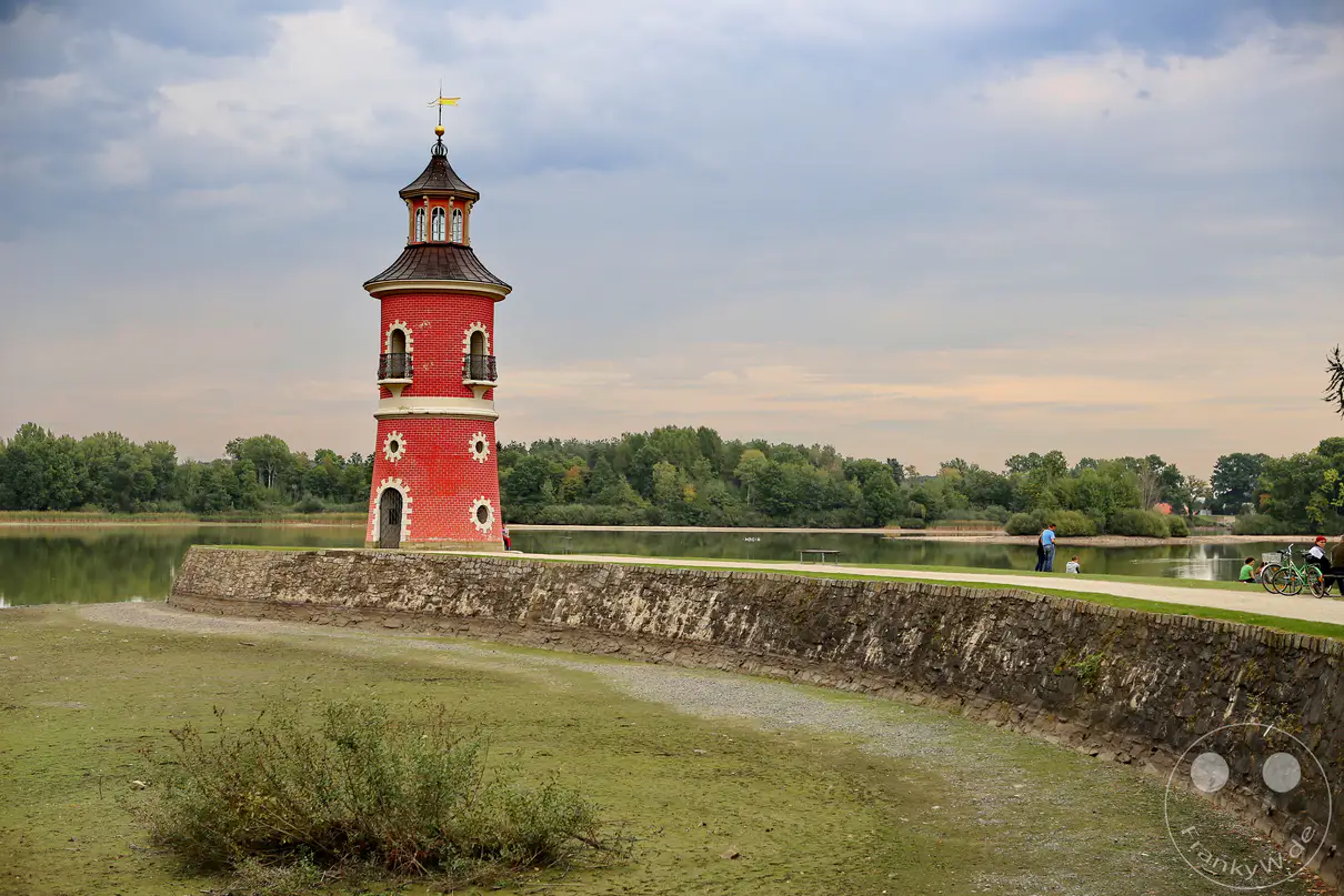 Der historische, rosafarbene Leuchtturm Moritzburg ragt am Ende einer steinernen Mole unter einem wolkenverhangenen Himmel empor. Das ruhige Gewässer des Großteichs umgibt die Anlage in Sachsen, Deutschland, während im Hintergrund eine dichte, grüne Waldkulisse die Szenerie einrahmt. Die gedämpften Farben und der weite Bildaufbau erzeugen eine friedliche, fast melancholische Stimmung an diesem außergewöhnlichen Ausflugsziel.