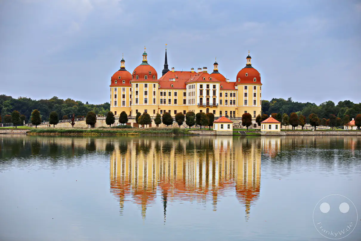 Schloss Moritzburg, ein prachtvolles Barockschloss in Deutschland, spiegelt sich an einem bewölkten Tag auf dem ruhigen Wasser des Schlossteiches. Die Architektur des Schlosses mit seinen markanten Kuppeln und der gelben Fassade hebt sich eindrucksvoll von der umgebenden grünen Baumkulisse ab. Die friedliche und majestätische Atmosphäre des Anblicks wird durch die symmetrische Reflexion im Wasser unterstrichen.