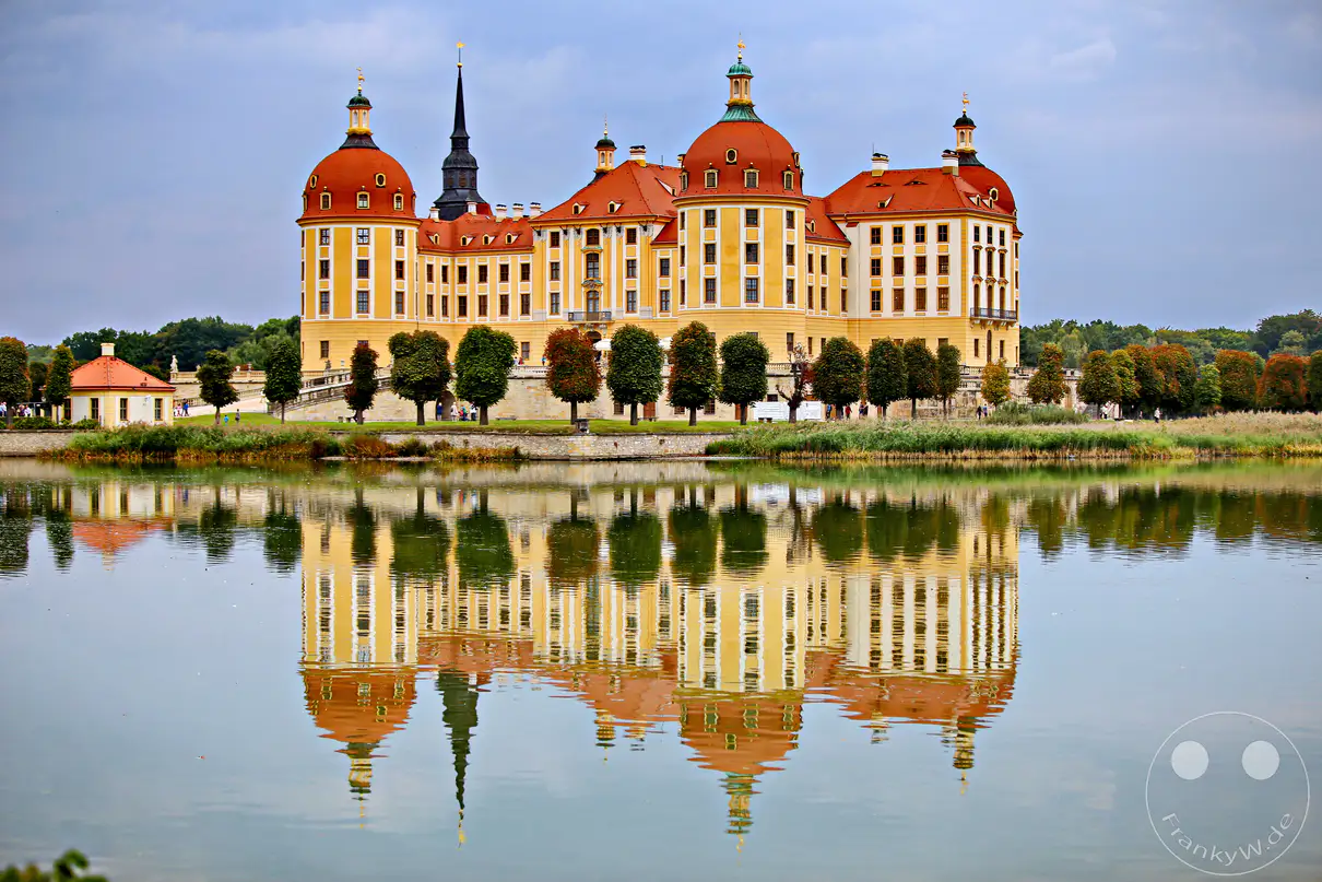 Schloss Moritzburg in Deutschland, nahe Moritzburg, präsentiert sich majestätisch auf einer Insel in einem malerischen See. Die symmetrische Spiegelung des barocken Bauwerks im ruhigen Wasser reflektiert die prachtvolle Architektur und die umgebende Natur. Eine friedliche und beeindruckende Szenerie, die die Schönheit des Schlosses und seiner Lage hervorhebt.